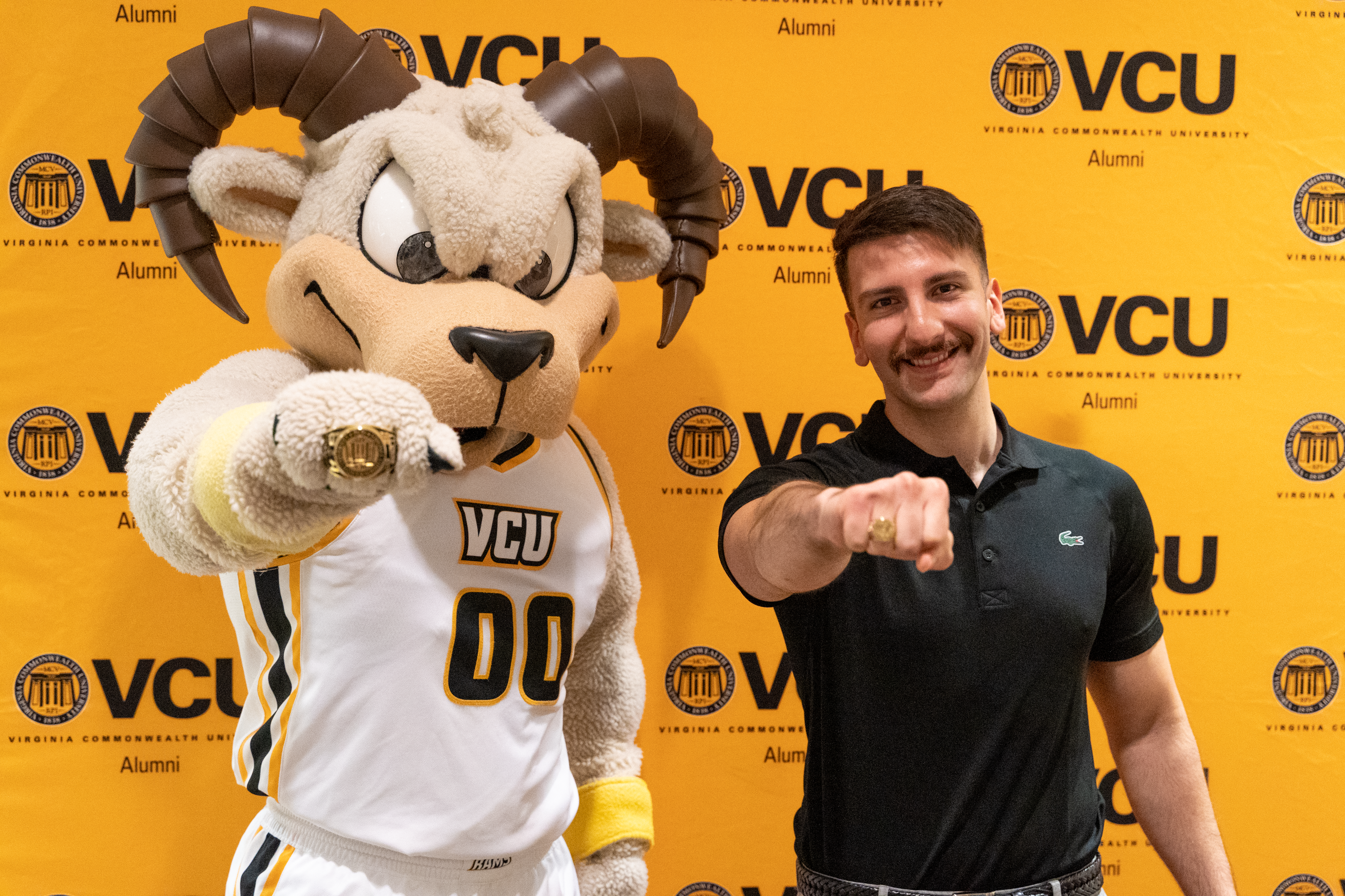 Rodney The Ram and a student showing off their VCU class rings in front of a gold backdrop